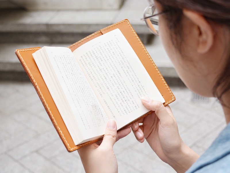 職人手作皮革厚革書套 (KB-4) M，淺棕色皮革，封面以車縫線裝飾，內含一本翻開的日文書籍。