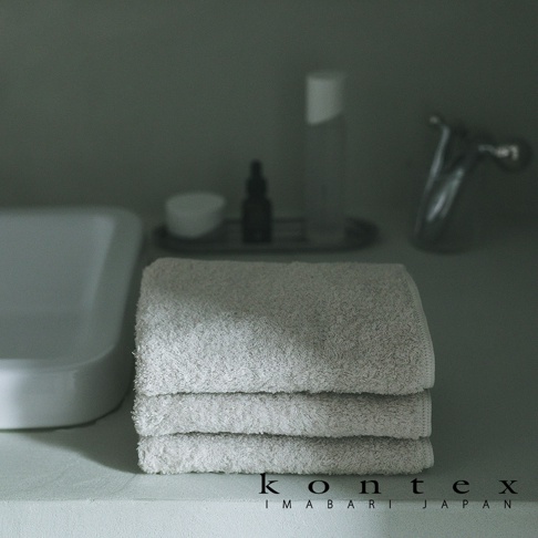 浴室洗手台上整齊堆疊著三條淺米灰色的 KONTEX 棉質毛巾，背景設有極簡風格的洗手盆與護膚瓶罐，整體呈現出沈穩且具有質感的日式生活氛圍。