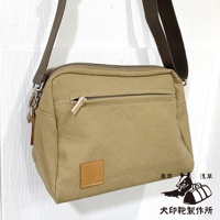 一個卡其色石洗帆布旅行側背包，配有深棕色帆布背帶，包包前方有一個拉鍊口袋，下方有一個方形皮革標誌，右下方有狗和行李的黑色圖案，以及日文文字。