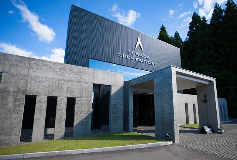 位於藍天白雲下方的諏訪田製作所開放工廠外觀，主體建築結合灰色清水混凝土牆面與黑色金屬浪板構造，大門上方懸掛白色蘇瓦達開放工廠英文字樣，周邊有草地與鋪設石塊的戶外空間，呈現簡約的現代建築風格。