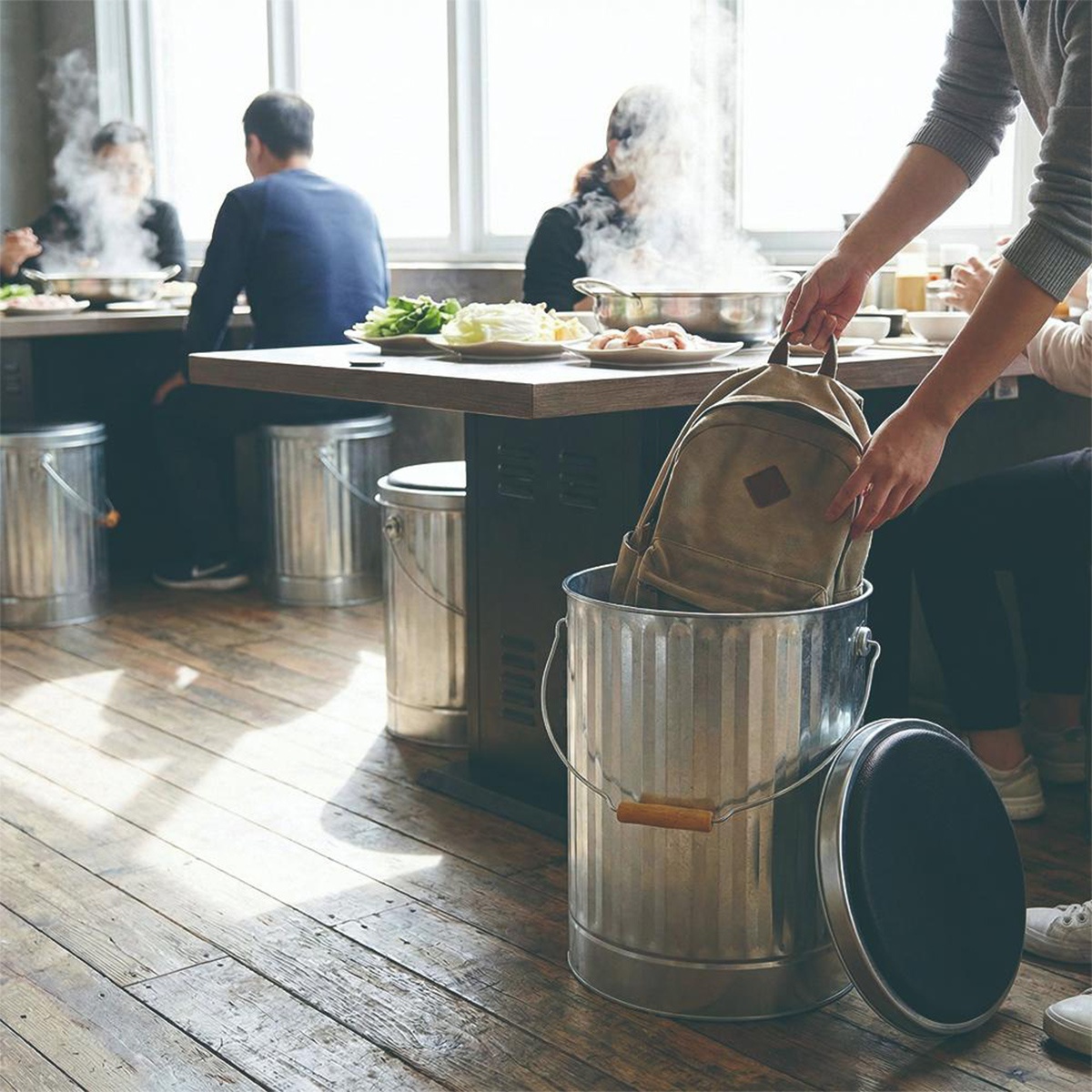 幾個人在室內用餐，其中一人將一個帆布背包放入銀色儲物桶內，旁邊還有一個蓋子。