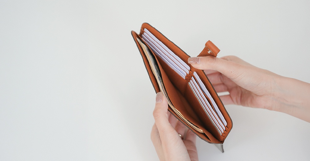 這張照片以俯視角度呈現一個敞開的 Atelier nuu 品牌棕色皮革雙折長夾，兩隻手分別輕扶著錢包兩側。錢包內部設有多個白色卡夾層與鈔票夾層，展現出俐落且簡潔的收納空間設計，整體色調溫潤且富有質感。