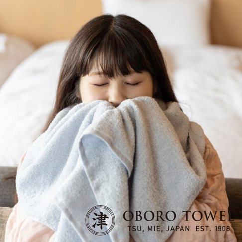 一位女子用淺藍色毛巾輕柔地觸碰臉頰，毛巾質地柔軟，帶有細緻的紋理，背景為模糊的臥室環境。