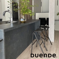 兩張黑色金屬框架的吧檯椅，椅面為黑色，椅腿呈細長交叉狀，擺放在深灰色石紋吧檯旁，地板為淺木紋。