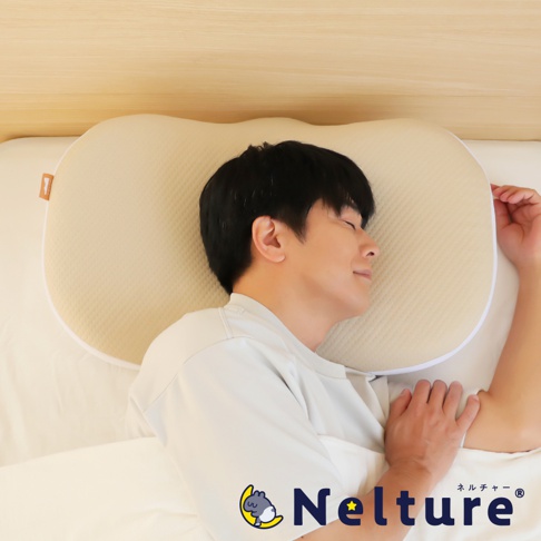 一名男子側臥在米色網狀織物製成的 NELUD 完美釋壓Q彈棉花糖枕上，枕頭呈腎形，邊緣飾有白色滾邊，枕頭上印有 NELTURE 品牌標誌。