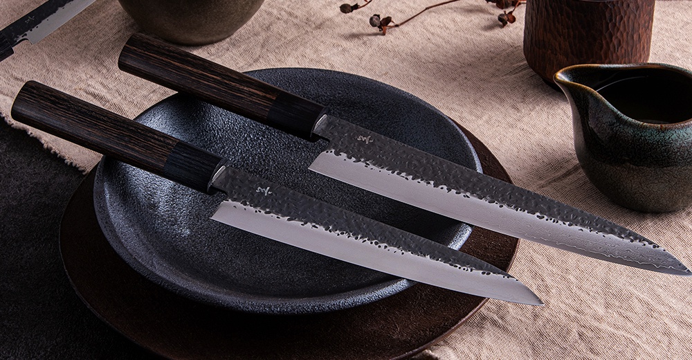 兩把玄 / Chef's knife 主廚刀，刀刃為鍛打紋路，呈現金屬光澤，刀柄為深棕色木質，放置於黑色盤中，背景為淺棕色布料。