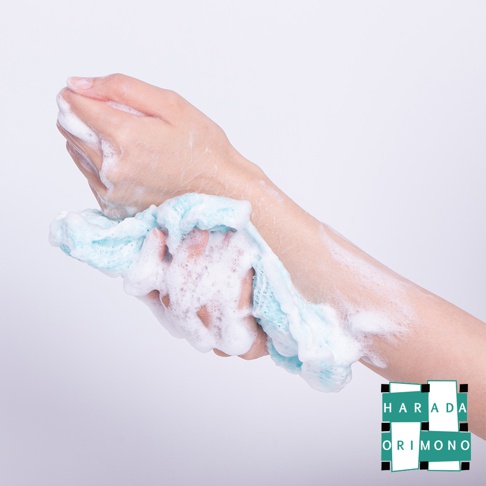 一隻手拿著淺藍色、網狀材質的泡泡洗澡巾，洗澡巾產生豐富的白色泡沫，覆蓋在手部和手臂上。