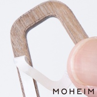 展示一個木製標示牌的局部特寫，邊緣有細緻的木紋，牌面為淺棕色。一隻手指正將背面的白色黏膠剝離，準備安裝。