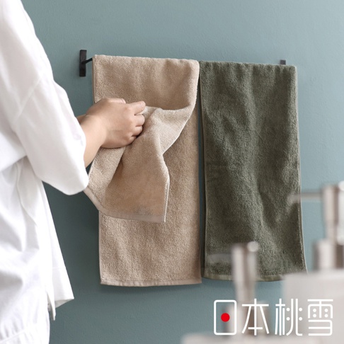一個人在浴室拿著一條米色毛巾，背景是掛在牆上的兩條毛巾，一條米色，一條軍綠色，右下角有日本桃雪的標誌。