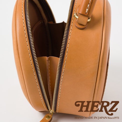 HERZ 職人手作皮革兩用圓形包，可見其精緻車縫細節與金屬拉鍊，品牌 LOGO 壓印於皮革上。