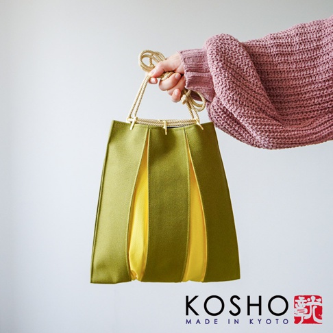 一名穿著粉色針織衫的女性手持京都KOSHO品牌的ougi帆布信玄袋，這款手提袋採用橄欖綠色與亮黃色拼接的褶皺設計，搭配米色編織繩提把，整體呈現簡約時尚的京都工藝風格。