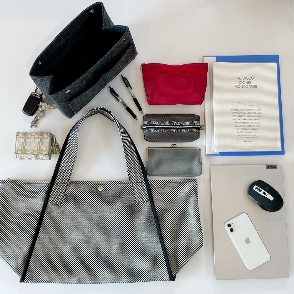 一個黑白格紋的帆布托特包，旁邊擺放著一個深灰色毛氈內袋、一個小巧的編織卡夾、兩支筆、一個紅色化妝包、一個灰色零錢包、一個文件夾、一台筆記型電腦、一個滑鼠及一支手機。