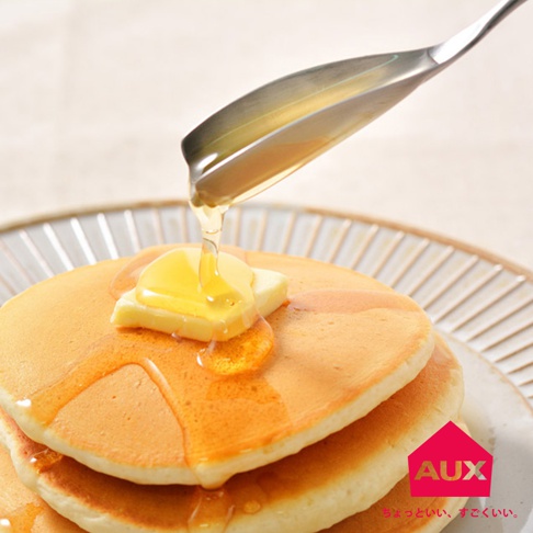 不鏽鋼蜂蜜匙，匙面盛著金黃色蜂蜜，正滴落在疊起的鬆餅和奶油上。