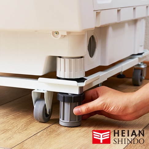 白色洗衣機下方放置著一個白色金屬可伸縮移動式平台，平台上裝有四個灰色滾輪和可調節高度的灰色底座，一隻手正在旋轉其中一個底座。