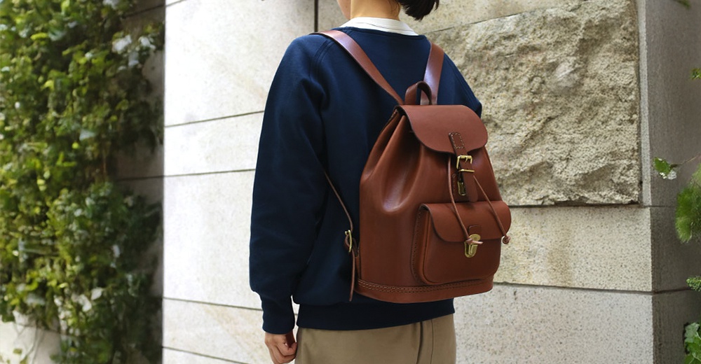 棕色皮革經典後背包，配有翻蓋、抽繩閉合、金屬扣環及前袋，由人體背負。