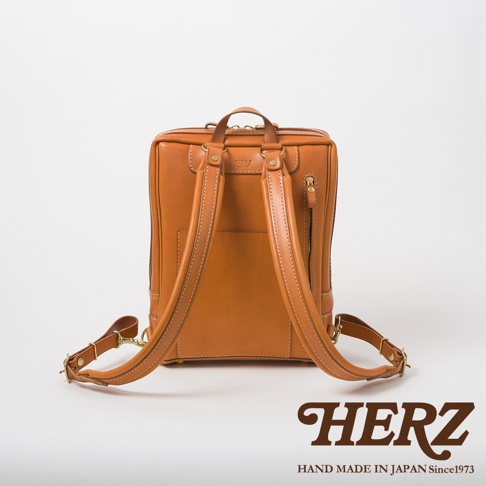 職人手作皮革軟皮商務後背包，展示背部，採用天然棕色皮革，搭配米白色縫線。背包頂部有提把，兩側有可調節的皮革肩帶，肩帶與背包主體以金屬釦件連接。背包側面有拉鍊口袋。