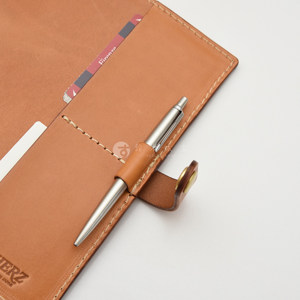 職人手作皮革A5手帳/記事本書套，採用天然皮革製作，呈現溫潤的淺棕色。書套內有筆套設計，可固定一支金屬筆，並有卡槽可放置證件或名片。車縫線細緻，展現皮革工藝。