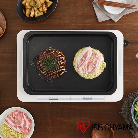 左右溫控電烤盤 WHP-012 頂部視角，黑色烤盤上放著兩個日式煎餅，左邊的灑有蔥花，淋上醬汁，右邊的煎餅鋪著切片豬肉。烤盤為白色外框，下方有兩個滑桿式溫控開關。