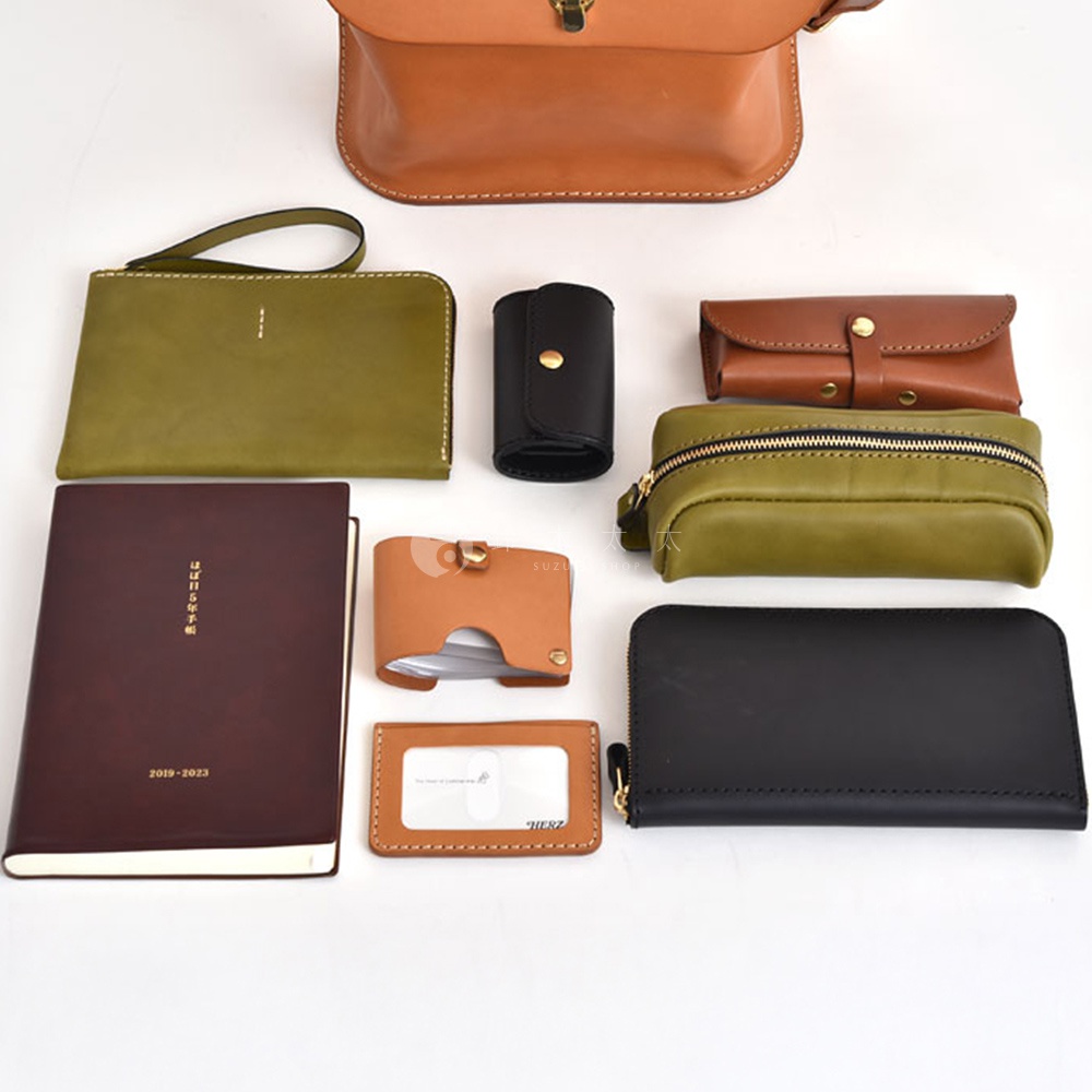 多款職人手作皮革配件，包含一個棕色翻蓋迷你後背包、一個綠色手拿包、一個黑色鑰匙包、一個棕色卡夾、一個綠色拉鍊筆袋、一本深棕色筆記本，以及一個棕色證件套。