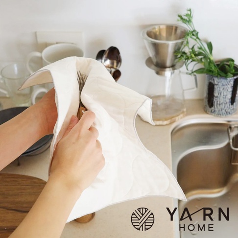 一雙手正在使用白色絎縫的廚房拭巾擦拭餐具，拭巾邊緣有灰色滾邊。背景為廚房水槽、咖啡濾杯與綠色植物。