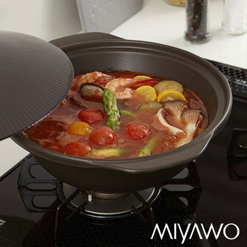 灰黑色陶土湯鍋，鍋內盛有豐富的湯料，包含鮮蝦、蛤蜊、蘆筍、小番茄、櫛瓜片及香菇等食材，湯鍋置於黑色爐架上，鍋蓋為深灰紋理狀。