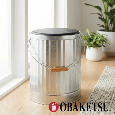 OBAKETSU 純銀色手工製作的鍍鋅鋼儲物凳，配有木製手柄和黑色坐墊，適合用於室內或戶外空間。