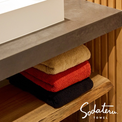 黑、紅、米色毛巾堆疊在木架上，上方是灰色檯面，右側有木條背景，右下角有 Sodateru TOWEL 的 Logo。