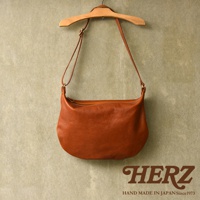 職人手作的駝色皮革單肩半月包，以自然垂墜的姿態掛在木質衣架上，包體飽滿，展現皮革的細緻紋理與光澤。