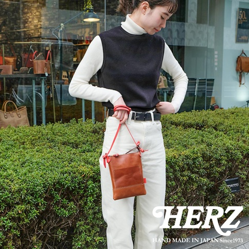 一位女性手持一個棕色皮革直式隨身單肩包，包包上有一個小口袋和紅色的皮革裝飾。