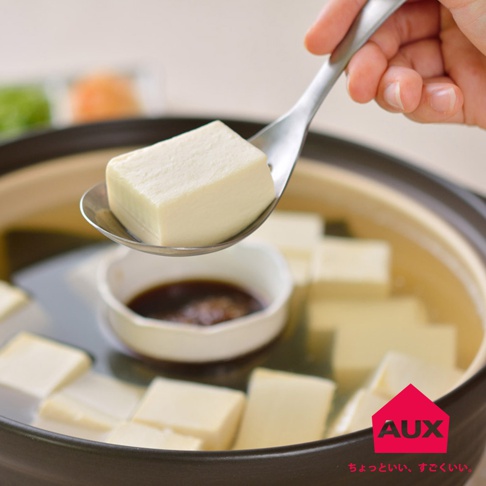 一只手拿著一個金屬湯匙，湯匙上盛著一塊白色的豆腐。湯匙下方是一個裝有醬汁的小白碗，置於一個裝滿湯水和豆腐塊的鍋中。