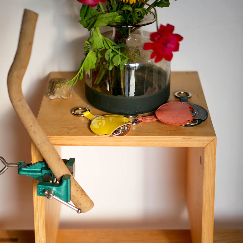 FABRIK 皮革圓型收納鑰匙圈，共有黃色、橘紅色和灰色三種顏色，展示於木製層架上，背景有鮮花與綠葉，鑰匙圈旁還有一個木製的彎曲握把被綠色鉗子夾住。