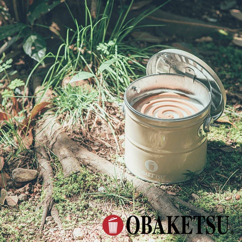 OBAKETSU手工製蚊香桶，裝有螺旋狀的蚊香，放置於佈滿青苔和落葉的地面上，周圍有綠草和樹根，蓋子被掀開放在一旁，上面有日文標示。