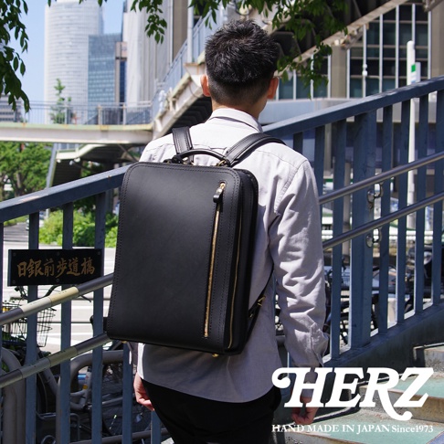 黑色皮革材質，方形設計的職人手作商務電腦後背包，配有金色拉鍊，由一位男士背在身上，背景為都市街景。