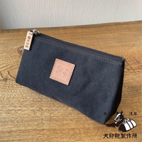 深藍色石洗帆布製成的小型收納包，包身有一個淺棕色皮革方塊標籤，上面壓印有圖案和文字。包包頂部有拉鍊，並有一個木質吊飾，上面刻有品牌標誌。