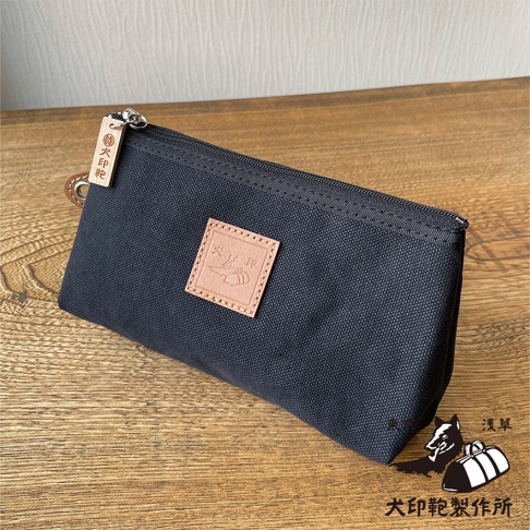 深藍色石洗帆布製成的小型收納包，包身有一個淺棕色皮革方塊標籤，上面壓印有圖案和文字。包包頂部有拉鍊，並有一個木質吊飾，上面刻有品牌標誌。