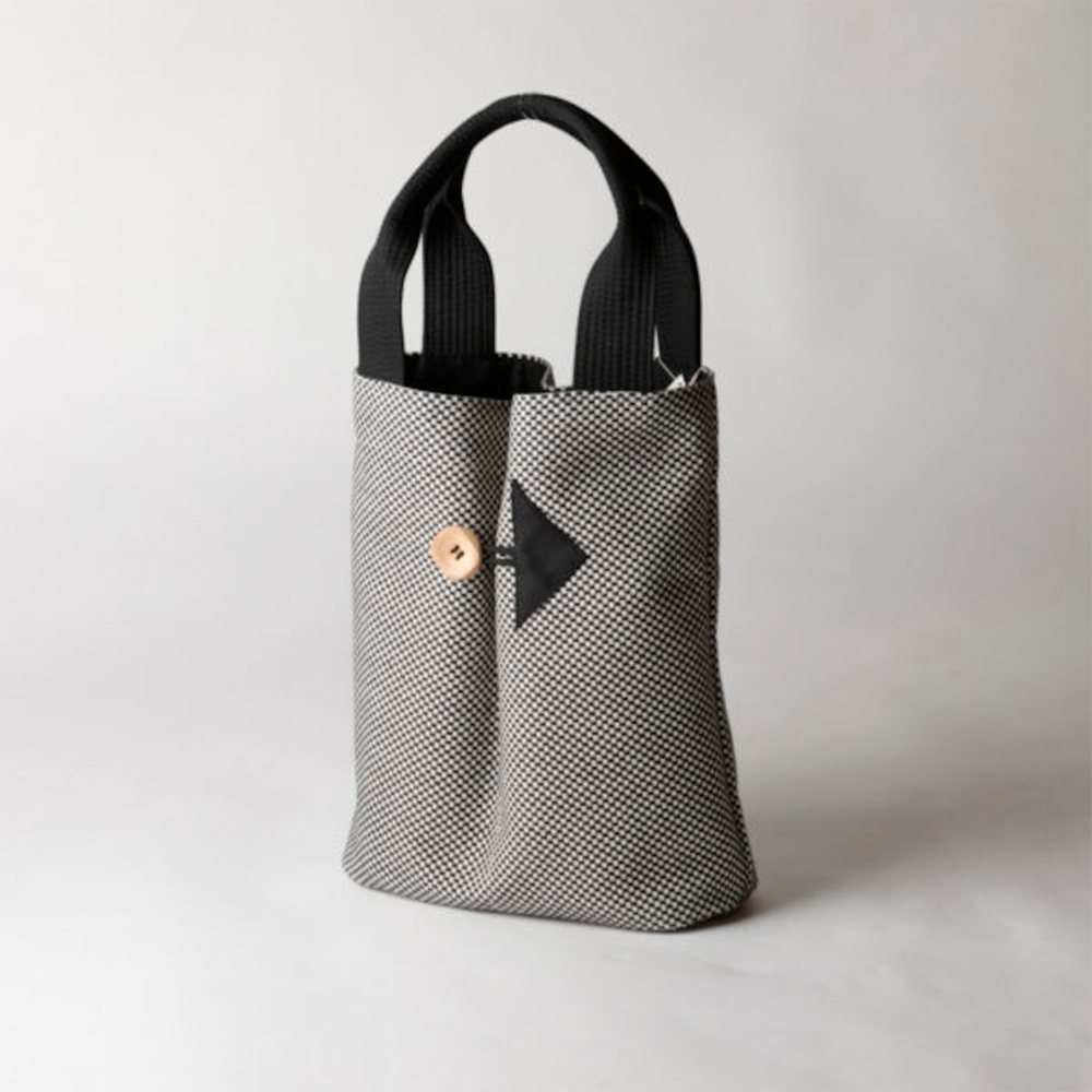 一款直式收納托特包，採用黑白棋盤格紋的三河木綿刺子織布料製成，配有黑色提把。包包中央有一個黑色三角形裝飾，並以一顆圓形木質鈕扣固定。