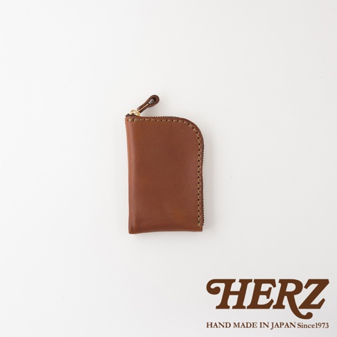 一款日本HERZ品牌職人手作的棕色皮革零錢包，表面呈現質感皮革紋理，邊緣採用縫線設計，頂部配有拉鍊與皮革拉環，右側邊緣圓潤，整體呈現簡約風格，右下角印有HERZ品牌標誌與產地資訊。