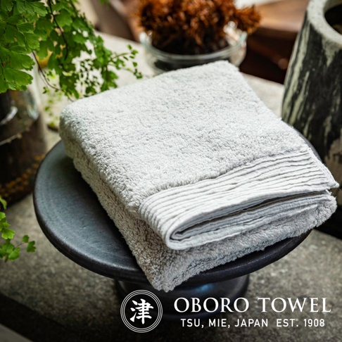 一張淺灰色毛巾，質地柔軟，邊緣有條紋裝飾，置於黑色圓形底座上。