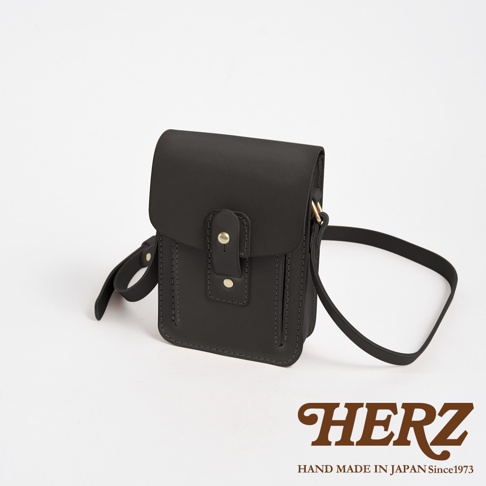 黑色皮革製成，方形翻蓋設計，帶有金屬鈕扣和縫線細節，可調節肩帶。