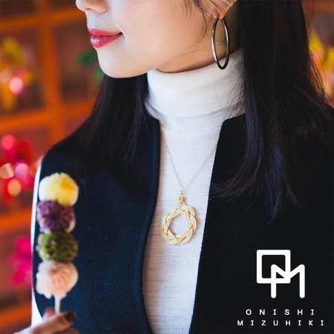 女性佩戴和風水引花結金絲項鍊，項鍊墜飾為金色編織水引，圓形設計，懸掛於金色細鍊上，搭配白色高領上衣和黑色外套。