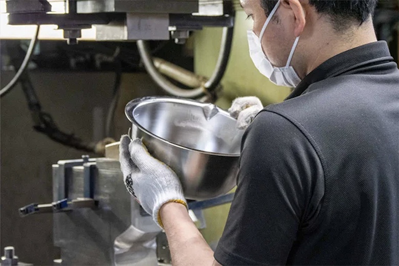 工人戴著手套及口罩，手持一個銀色不鏽鋼濾水網與刻度料理盆。