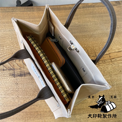 犬印純棉帆布迷你托特包，米白色帆布材質，深棕色提把，包內裝有筆記本、手機和皮夾，包蓋處有金屬鎖扣，右下角有品牌標誌。