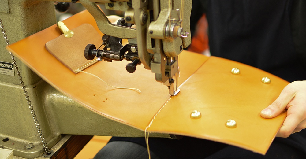 職人使用工業縫紉機縫製原色皮革，皮革上固定著金屬釦，呈現製作過程的細節。