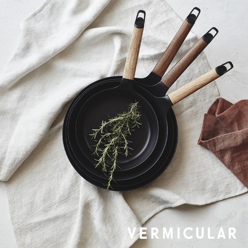 日本製鑄鐵平底深鍋，黑色鍋身，配有木質手柄，鍋內飾有迷迭香，置於白色亞麻布上。