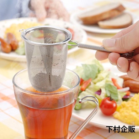 手持不鏽鋼濾茶網，網篩呈圓柱狀，網孔細密，裝有茶葉，置於裝有茶飲的玻璃杯上方，背景為早餐餐點。