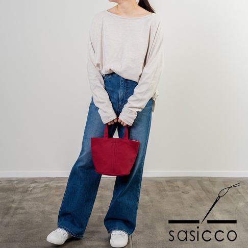 一位女性手持紅色三河木綿刺子織圓底水桶包，搭配淺色長袖上衣與牛仔寬褲。