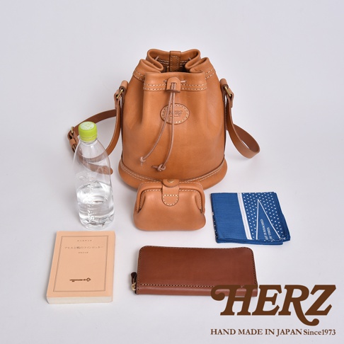 職人手作皮革束口單肩包，採用質感棕色皮革製作，設計經典。包身束口設計，配有可調節肩帶。周邊擺放著同色系皮革零錢包、長夾、一本淺棕色書籍、一瓶礦泉水及一塊藍白印花手帕，展示產品容量與搭配性。