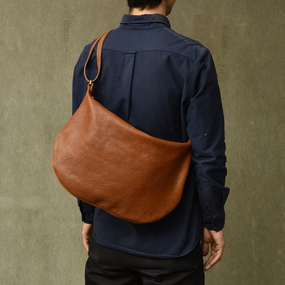 棕色皮革製的單肩半月包，斜挎於身穿深藍色襯衫的模特兒身上。