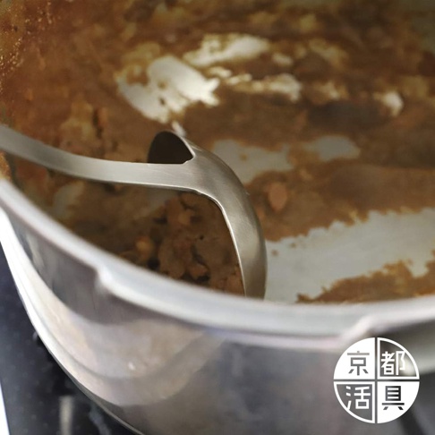 不鏽鋼萬用料理大勺的勺頭部分，置於裝有褐色食物殘渣的鍋中，勺柄為銀色金屬材質，鍋身為銀色金屬。