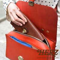 HERZ 品牌紅色皮革多功能收納包，一只手正在將長夾放入包內，包內可見藍色小物，下方有 HERZ 品牌標誌與 Handmade in Japan Since 1973 字樣